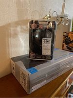 Wireless headphones in sealed plastic packaging showing branding and features like noise reduction, volume control, transmitter, and 50ft range cable.