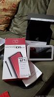 Nintendo DS Lite handheld console in red with its original box and manual alongside Amazon Echo Show device and its box on a couch.