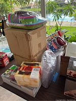 Boxed and bagged colorful plastic plates and bowls on top of a cardboard box with other boxed kitchen and party items nearby.