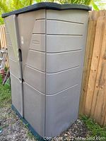 Side view of the tall beige Rubbermaid outdoor storage container placed next to a wooden fence with visible vegetation.