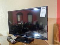 Samsung TV on black stand with remote in front. TV screen is off showing reflections of room.