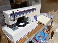 Front and side view of Husqvarna Viking sewing machine with power cord on work table, alongside accessories and fiber bundles.