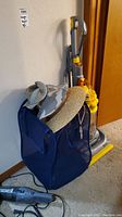 Dyson upright vacuum cleaner, Bissell handheld vacuum, straw hat, other hats, and dark blue folding basket hamper on carpeted floor near wooden door and wall.
