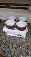 Four white ceramic bowls set in brown wicker baskets on a patterned cloth on a kitchen counter.