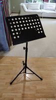 Front view of the adjustable black metal music stand on a hardwood floor with a carpet and couch in background.