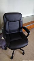 Front view of black leather-look office chair showing padded seat and backrest, fixed armrests, and caster wheels.