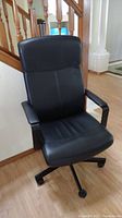 Front-left angle view showing tall cushioned backrest, seat, and armrests of the black leather-look office chair on caster base.
