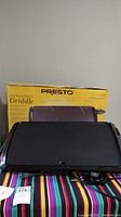 Photo showing the electric griddle placed on a colorful tablecloth with the original yellow Presto box behind it, displaying the front and top view of the griddle's cooking surface and cool touch handle.