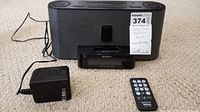 Black Sony clock radio with speaker and iPod dock, power cord, and remote placed on carpeted floor. Speaker finish shows cracks and wear.