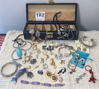 Top-down view of assortments of jewelry items including bracelets, earrings, necklaces, keychains, buttons, and brooches displayed in front of an open black jewelry box.