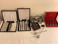 Photo of entire lot arranged in three parts: two black boxes of utensils on left, silver plated accessories and snuff box in center and right with red box of spoons.
