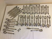 Photo showing the full set of flatware arranged on a white cloth. Includes dinner forks, knives with black handles, spoons, dessert forks, dessert/tea spoons, a spreader, and a sugar spoon.