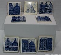 Eight ceramic tiles/coasters arranged on white risers. Four loose tiles with blue architectural building designs, four tiles sealed in original KLM business class blue and white boxes.