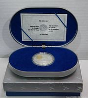 Oval blue velvet-lined display case containing silver coin in protective capsule, with certificate inside lid, resting on outer box with aircraft motif.