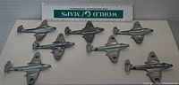 Seven diecast Gloster Meteor model airplanes displayed with a measurement ruler for scale, showing front and top views revealing silver paint finish and roundel decals. Various conditions, some paint wear visible.