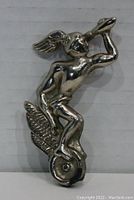Front view of a shiny silver-tone metal figure of a winged male rider sitting on a winged wheel, holding an elongate object to the mouth, against a plain background.