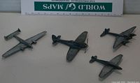 Four metal toy planes arranged on a light surface with a ruler and World War maps booklet for scale and context. Shows top view and relative sizes.