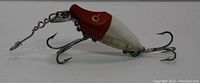 Side view of the fishing lure showing red head, white body, treble hooks and attached chain and swivel.