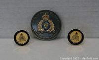 Photo showing one pair of gold plated RCMP cufflinks with cream and gold crests and black edging, paired with a black RCMP forensic science service challenge coin with a detailed crest in the center.