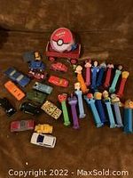 Photo showing a red plastic case with Pokemon playing cards and an assortment of colorful Pez dispensers and toy cars scattered on a dark surface.