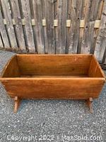 Front and side view of primitive wood cradle on pavement outdoors with wooden fence background, showing rectangular body and rounded legs supporting the cradle.