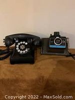 Northern Electric black rotary dial telephone and Polaroid instant camera side by side on brown surface.