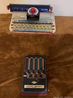Junior Marx Typewriter on top with the Wolverine Adding Machine below on a brown fabric surface.