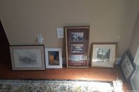 Five art prints displayed at floor level, four framed, one unframed leaning against wall on wood floor