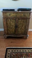 Front view of wooden painted dinner cabinet with floral design, single drawer, double-door lower cabinet, placed on hardwood flooring with a patterned rug in front.