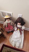 Two porcelain dolls positioned sitting and standing on wood floor against wall with electrical outlets and vent visible.
