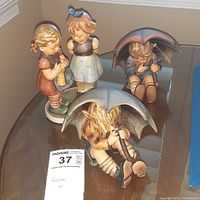 Set of three Goebel figurines on glass table: boy with umbrella sitting, two girls standing together, girl with umbrella standing.