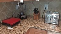 Photo showing the four kitchen items on a granite kitchen counter: red George Foreman grill, black KitchenAid blender, stainless steel Cuisinart toaster, and Wusthof knives in wooden block.