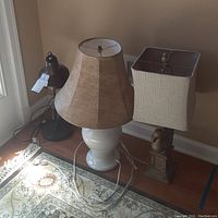 Three lamps shown together on a floor with tag on black desk lamp. Includes white ceramic base lamp with tall beige shade and bird-themed lamp with square shade.