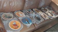 Set of 12 Norman Rockwell Christmas plates displayed on couch with bubble wrap, showing multiple designs