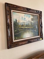Oil painting hung on wall showing landscape scene with pond, trees, and mountains under a cloudy sky, set in a carved wooden frame.