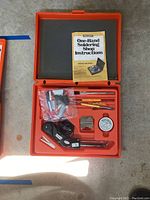 Opened orange plastic case showing One-Hand Soldering Shop Kit including soldering iron, tips, cleaning paste, and instruction manual