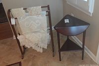Wooden towel rack holding crocheted towels beside a triangular drop leaf wooden table with a lower shelf, both with a dark finish