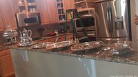 Wide view showing all silver plated serving items arranged on kitchen counter, including coffee service, trays, chafing dish, and butter dish.