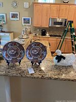 Photo of two Asian decorative plates with intricate floral and bird designs displayed on black stands, alongside a white ceramic duck-shaped soup tureen with a green ribbon and ladle on a kitchen counter.