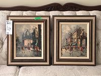 Pair of framed Parisian street scene oil paintings on couch with tags visible