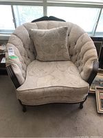 View of wood-framed bucket armchair with matching square cushion, beige brocade fabric upholstery, and button tufting, positioned near window.