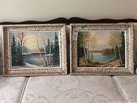 Two framed landscape oil paintings on board displayed side by side on a white quilted surface; both have textured cream-colored frames and show natural scenes with trees and waterways.