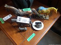 Several cast metal horse figurines along with cast metal goat lighter, Wedgwood ashtray, and metal bell without clapper on wooden table.