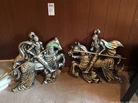 Two cast metal wall plaques of jousting knights on horses, showing size difference and details of armor and horses