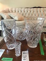 Four clear crystal pieces on table including two vases, a bowl, and a small bud vase, shown with wood table and beige tufted couch backdrop.