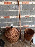 Wide shot showing all three items: demijohn in wicker basket, wooden barrel stand, and garden edger leaning against wall