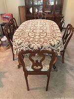 Dining table set covered with a white lace tablecloth, showing six wooden chairs with carved backs around it.
