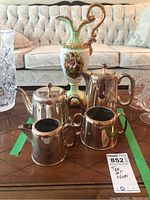 Photo showing silverplated tea set consisting of teapot, cream, and sugar bowl plus the ceramic ewer with floral design on a wood table.