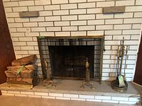 Photo showing the fireplace with brass andirons, brass log grate holding firewood, metal mesh fireplace screen, and brass tool stand with tools.