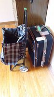 Photo showing the plaid fabric folding cart with metal frame and black and brown wheeled luggage side by side on a hardwood floor.